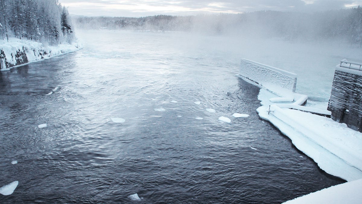 En vinterälv omgiven av snö och is, med en snötäckt damm på höger sida.